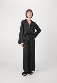 Svart satinpyjamas-set som består av en knapp-up skjorta med krage och långa ärmar, tillsammans med vida byxor och lurviga svarta tofflor.