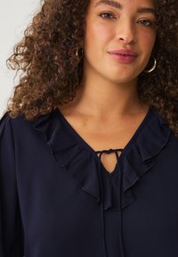Navy blouse met een ruffles V-hals en een striksluiting. Gemaakt van een gladde stof met een zachte textuur. Gouden ringoorbellen zichtbaar.