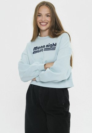 Junge Frau mit langen braunen Haaren, die ein hellblaues Sweatshirt mit dem Text "moon night music" und schwarze hochgeschnittene Hosen trägt, lächelt mit verschränkten Armen.