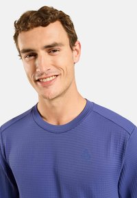 Lila Sportshirt mit strukturiertem Muster, rundem Halsauschnitt und Logodetail auf der Brust. Kurzarm und taillierte Passform.