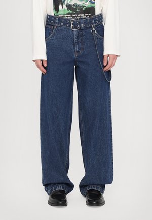 Wide-leg denim jeans in dark blue, featuring a large belt with metal eyelets and a chain detail. Textured fabric with visible stitching.