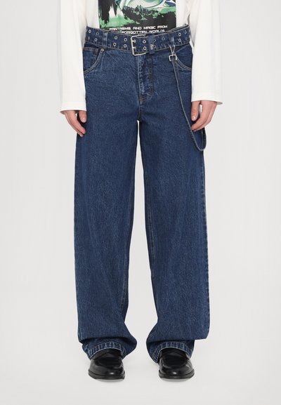 Jeans de denim de pierna ancha en azul oscuro, con un gran cinturón con ojetes metálicos y un detalle de cadena. Tejido texturizado con costuras visibles.