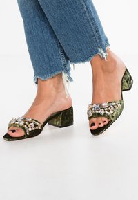 Vihreät samettiset liukusandaalit, joissa on lohkarekorko, koristeena isot kirkkaat ja meripihkanväriset jalokivet, avotopainos ja repaleiset farkut.