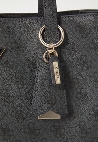 Crna tote torba s teksturom koja uključuje zlatne hardverske detalje, oznaku s logotipom i trokutasti naglasak. Materijal ima suptilnu uzorkovanu završnu obradu.