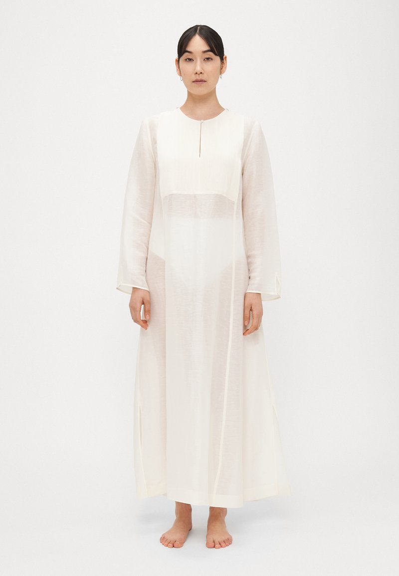 Ελαφρύ, κρεμ φόρεμα με μακριά μανίκια, στρογγυλή �λαιμόκοψη και πλαϊνές σχισμές. Διακοσμείται με υφασμάτινη εφαρμοστή λωρίδα στο μπούστο.