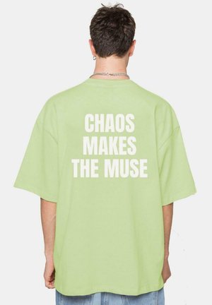 Camiseta de gran tamaño en color verde claro con cuello redondo; presenta un texto blanco en la espalda: "EL CAOS HACE LA MUSA." Textura de algodón suave.