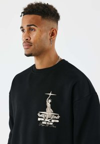 Jonge man met kort, krullend haar draagt een zwart sweatshirt met een afbeelding van het Vrijheidsbeeld en de tekst "Change The Game" aan de voorkant.