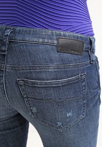 Jeans de mezclilla azul oscuro con un parche de cuero negro, diseño de cinco bolsillos y sutiles acentos desgastados en los bordes de los bolsillos traseros.