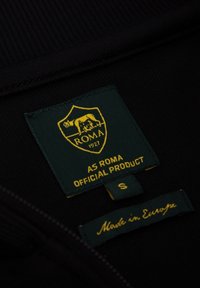 Etichetta nera con uno scudo verde, un emblema giallo e il testo: "PRODOTTO UFFICIALE AS ROMA," taglia S, e "Fatto in Europa."