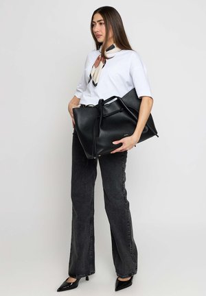 Donna con top bianco, jeans neri e tacchi a punta tiene una grande borsa tote in pelle nera, indossa una sciarpa fantasia al collo.