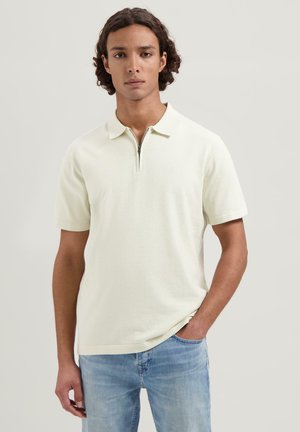 Hellescremefarbener Poloshirt mit Reißverschluss und kurzen Ärmeln, kombiniert mit hellblauen Jeans. Die Textur wirkt glatt und weich.