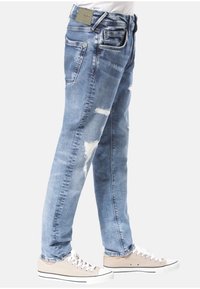 Světle modré džíny z denimu s distressed detaily, včetně roztržení, rovný střih nohavic a látková nášivka na zadním pásu. Nosené s béžovými teniskami.