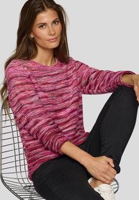 Rosa-rot gestreifter Pullover mit rundem Ausschnitt und langen Ärmeln, kombiniert mit schwarzen Hosen und weißen Turnschuhen. Weiche Stricktextur.