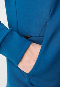 Tessuto in felpa blu con tasca con zip, polsini texture e finitura liscia sulla parte del corpo.