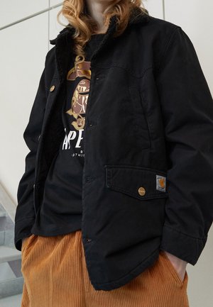 Persona con chaqueta negra Carhartt sobre una camiseta negra con estampado y pantalones de pana marrones, de pie en el interior cerca de unas escaleras.