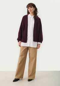 Cardigan bordeaux con una texture pelosa, apertura frontale e spalle scese, indossato sopra una camicia bianca con bottoni e pantaloni beige a gamba larga. Scarpe nere.