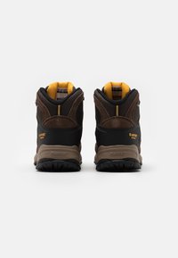 Brune vandrestøvler med sort mesh og ruskind, der har gule detaljer og strukturerede gummisåler for godt greb. Bagfra ses branding og detaljer.