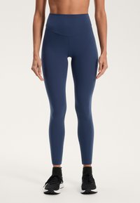 Sötétkék, magas derekú leggings sima textúrával, karcsú fazonnal és láthatatlan varrásokkal. Fekete sportcipővel viselve.