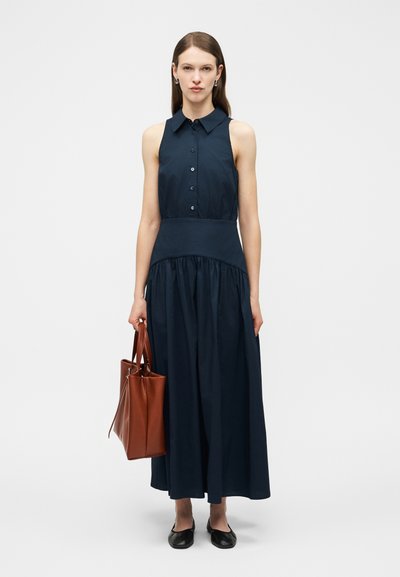 Femme en robe sans manches bleu marine tenant un sac fourre-tout en cuir marron, portant des ballerines noires, debout devant un fond clair uni.