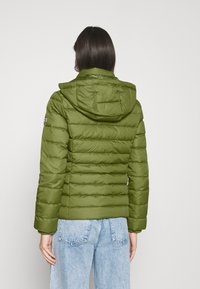 Mujer vestida con una chaqueta acolchada con capucha de color verde oliva y unos vaqueros de cintura alta en azul claro, vista desde atrás contra un fondo liso.