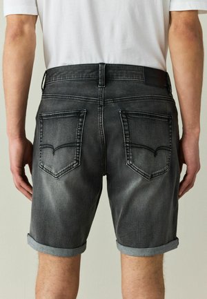 Mann trägt ausgewaschene schwarze Denim-Shorts mit aufgerollten Säumen und zwei Gesäßtaschen, von hinten vom Saum bis zur Mitte des Oberschenkels gezeigt.