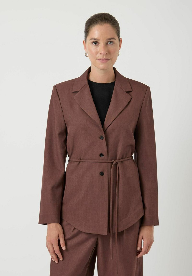 Vrouw in een bruine blazer met knopen en riem over een zwarte top, gecombineerd met bijpassende bruine broek, staand tegen een effen achtergrond.