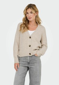 Cardigan de punto beige con escote en V, cuatro botones marrones, puños acanalados y corte relajado, combinado con jeans grises.