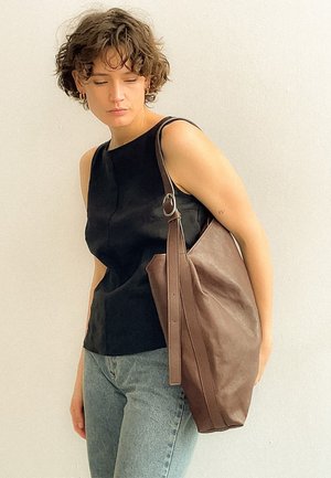 Mujer con cabello corto y rizado, vestida con una camiseta negra sin mangas y vaqueros, llevando un bolso grande de cuero marrón al hombro, frente a una pared lisa.