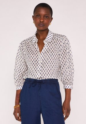 Femme portant un chemisier blanc à motifs de feuilles bleues et un pantalon bleu avec cordon de serrage, debout devant un fond clair uni.