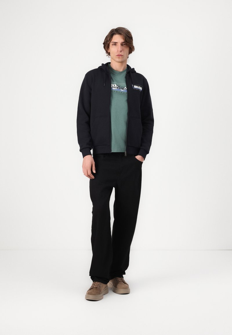 Młody mężczyzna w czarnym hoodie na zamek, zielonej koszulce z grafiką, czarnych spodniach i beżowych zamszowych sneakersach stoi na tle białej ściany.