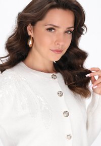 Cardigan alb tricotat cu închidere cu nasturi, decorat cu modele de frunze cu paiete. Prezintă nasturi rotunzi și lucioși cu un accent metalic.