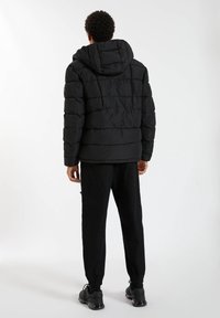 Personne debout, de dos, portant une doudoune noire avec capuche, un pantalon noir et des baskets noires sur un fond blanc.