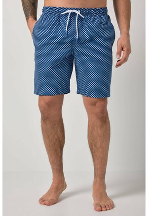 Mężczyzna w niebieskich i białych wzorzystych kąpielówkach z białym sznurkiem, stojący boso na jasnym tle.