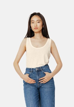 Femme aux longs cheveux foncés portant un haut sans manches crème et un jean bleu taille haute, mains posées sur les hanches, expression neutre.