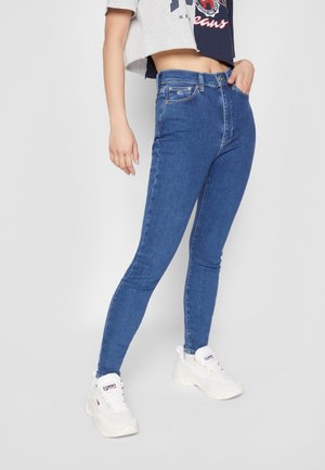 Osoba nosząca niebieskie skinny jeans z wysokim stanem, krótki dwukolorowy top i białe masywne sneakersy, stojąca na jednolitym tle.