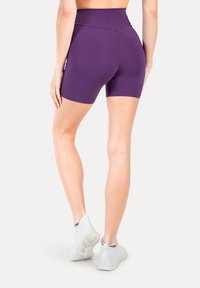 Shorts viola a vita alta realizzati in materiale elastico, caratterizzati da un design senza cuciture e una texture liscia, abbinati a scarpe da ginnastica bianche.