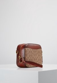 Ruskea nahkainen olkalaukku, jossa on vaaleanbeige canvaskangas etupuolella ja kuvioitu design. Kultaiset metalliosat ja vetoketjusuljin.