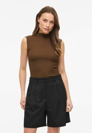 Jeune femme aux longs cheveux bruns portant un haut marron sans manches et un short noir taille haute cintré, debout devant un fond uni.