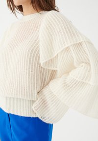 Maglione bianco lavorato a maglia con una texture a coste e ampie maniche a strati. L'orlo inferiore è aderente, in contrasto con il design ampio della parte superiore.