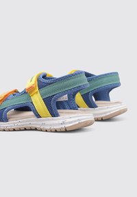Sandalias coloridas con tejido de malla en azul, verde y amarillo, con correas de Velcro, una suela texturizada y una plataforma blanca moteada.