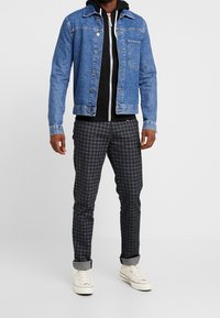 Uomo che indossa una giacca di jeans blu sopra un hoodie nero con zip, pantaloni a quadri grigi arrotolati alla caviglia e sneakers bianche alte.