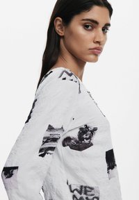 Cămașă albă cu mânecă lungă, prezentând imprimeuri foto alb-negru, material texturat cu un aspect ușor crispat și guler rotund.