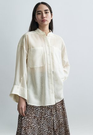 Chemise boutonnée crème clair à rayures verticales avec deux poches, portée sur une jupe imprimée animalier marron et blanc. Tissu doux et transparent.
