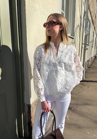 Valkoinen pitsipaita, jossa on pitkät hihat, kaulukseton muotoilu ja monimutkaiset kuviot. Yhdistetty valkoisiin farkkuihin ja tummanruskeaan käsilaukkuun.