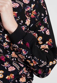 Sort stof med blomstermønster i orange, gule og hvide blomster. Manchetter med elastisk sort ribbet tekstur.