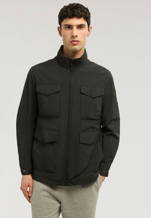 Mann trägt eine schwarze Jacke mit Reißverschluss und vier Fronttaschen, steht vor einem schlichten hellen Hintergrund und blickt nach vorne.