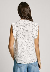 Blusa bianca senza maniche con pois blu navy, spalle arricciate e tessuto testurizzato. Abbinata a jeans blu a vita alta.