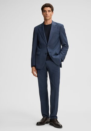 Granatowy garnitur z teksturowanym wzorem, jednorzędowa marynarka i spodnie bez zaszewek, połączone z czarną koszulą z okrągłym dekoltem i ciemnymi butami.