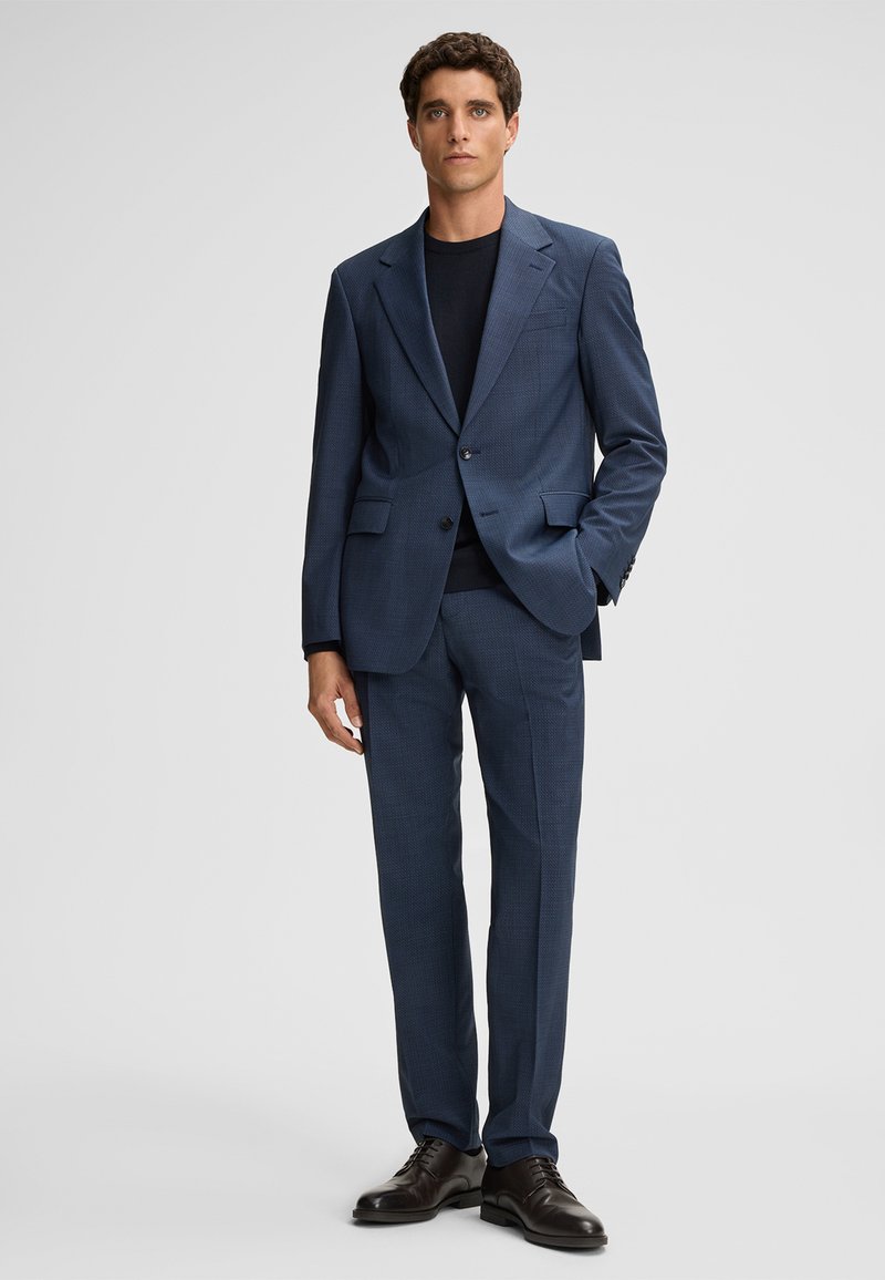 Dunkelblauer Anzug mit strukturiertem Muster, einreihige Jacke und gerade geschnittene Hosen, kombiniert mit einem schwarzen Rundhalshemd und dunklen Schuhen.