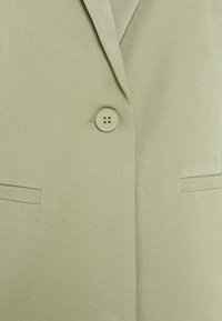 Blazer verde claro con una tela texturizada, que presenta un único botón ligeramente contrastante y dos bolsillos frontales. Ajuste entallado.